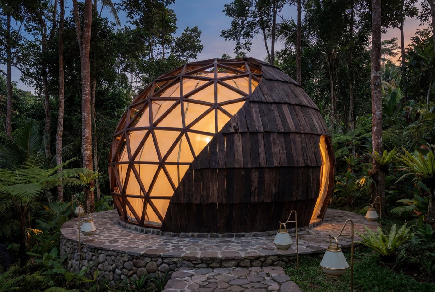 A geodesic sound-healing dome in ironwood and timber shingle, glowing warm at dusk on a stone podium in the jungle